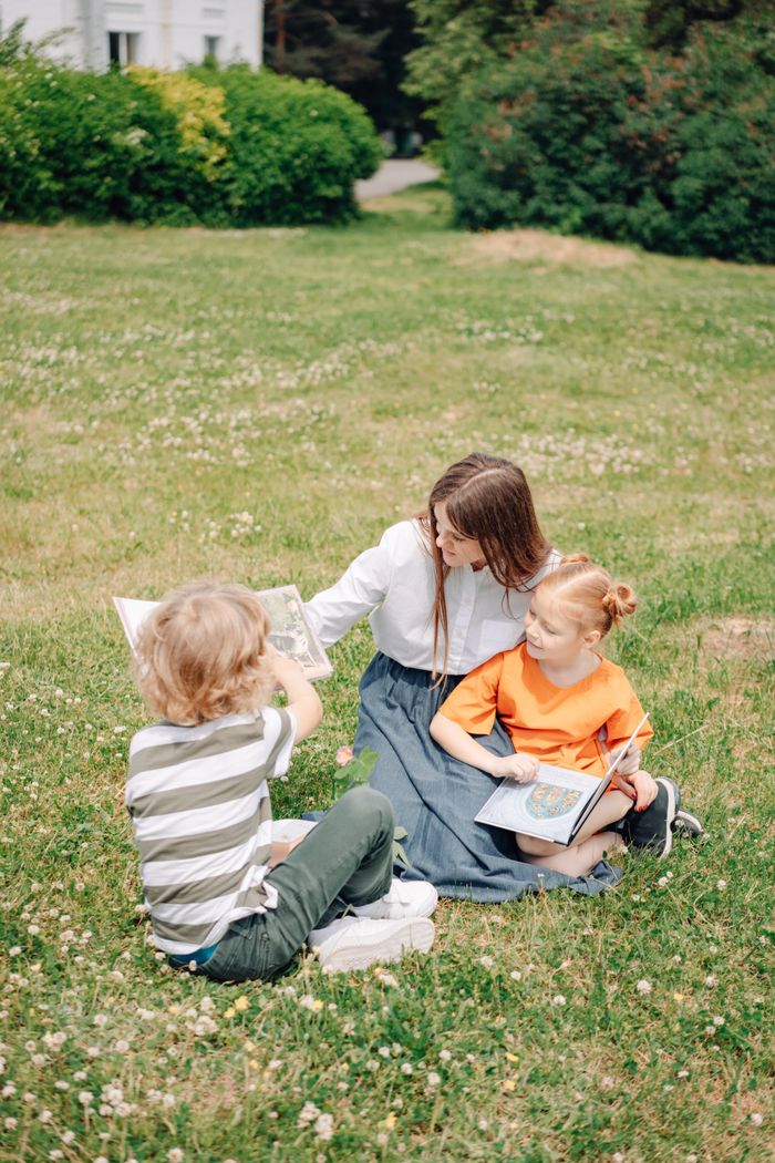 A teacher reads storybooks to children while sitting on lush green grass outdoors, fostering learning.